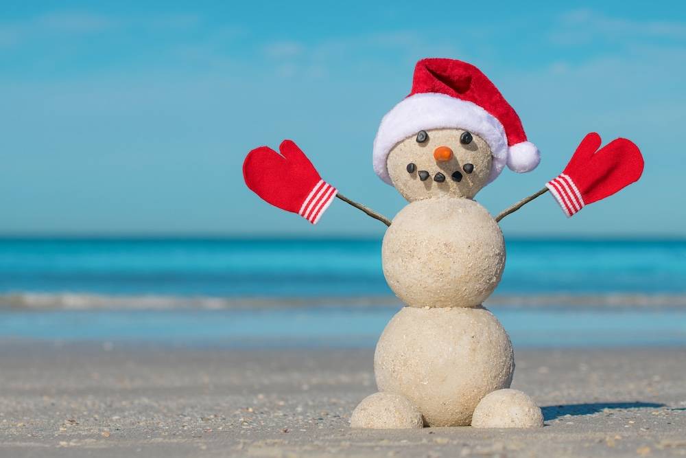 A sandcastle snowman with red mittens and a Christmas hat with the Atlantic Ocean in the background